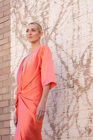 A woman modeling a coral ALEX KIMONO BY ONE FELL SWOOP with elbow-length sleeves, standing against a split brick and painted brick wall background.