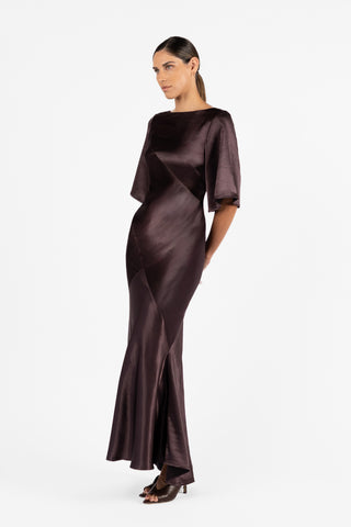 A side view of the Moni Gown in Aubergine by One Fell Swoop Moni Gown as it showcases a sleek bateau neckline, architectural bias-cut panelling, and sophisticated elbow-length sleeves.