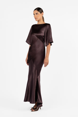 The One Fell Swoop Moni Gown in Aubergine showcases a sleek bateau neckline, architectural bias-cut panelling, and sophisticated elbow-length sleeves. Cut from luxurious plum satin with a radiant finish.