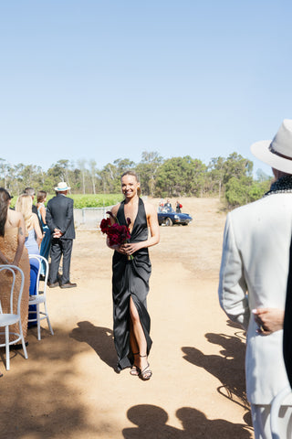Bridesmaid walking down the aisle at an outdoor wedding wearing the One Fell Swoop Talia Dress in Obsidian, holding a red floral bouquet