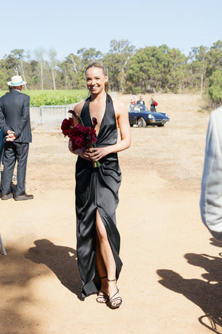Bridesmaid walking the aisle at an outdoor vineyard wedding wearing the One Fell Swoop Talia Dress in Obsidian — black satin halter maxi with twist knot waist detail