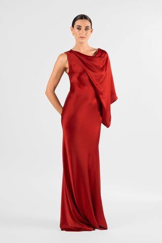 One Fell Swoop Glinch Gown in Carmine, front view with one hand tucked behind the hip, showing the deep red satin cowl neckline and the asymmetric draped panel that falls from the shoulder across the chest, with the sleeveless bodice fitted close to the body.