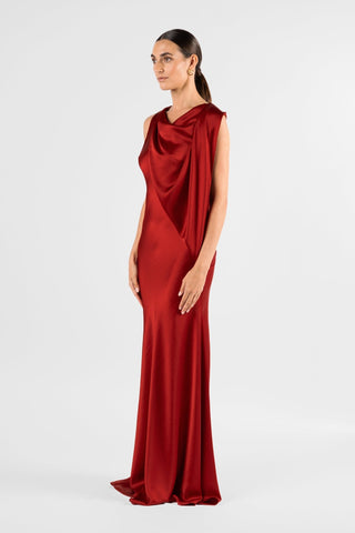 One Fell Swoop Glinch Gown in Carmine, second side three-quarter view in deep red liquid satin, showing the soft cowl at the neckline, the drape falling in a long diagonal across the front of the bodice and the narrow, bias-cut column skirt reaching the floor.