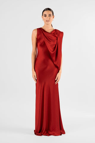 One Fell Swoop Glinch Gown in Carmine, straight-on front view standing with arms relaxed, showing the deep red liquid satin gown with a soft cowl neckline, a draped panel falling from one shoulder across the bodice, and a bias-cut skirt that widens gently to the floor.
