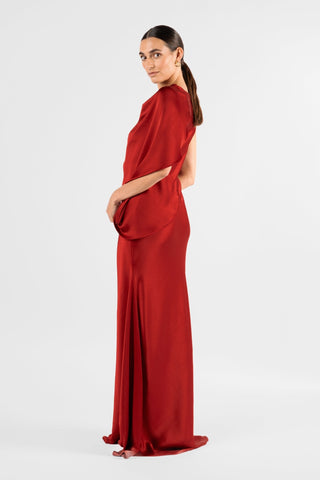 One Fell Swoop Glinch Gown in Carmine, three-quarter back view with the hand resting at the hip, lifting the draped wing of satin at the back shoulder, with the deep red bias-cut skirt flowing to a floor-length hem.