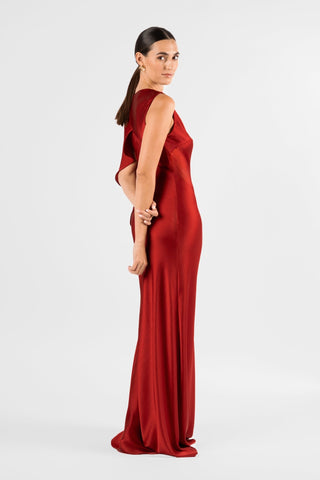 One Fell Swoop Glinch Gown in Carmine, model turning to look back over her shoulder, showing the draped cape-like panel at the back shoulder, the sleeveless fitted bodice and the long satin skirt falling smoothly from the waist in a deep red sheen.