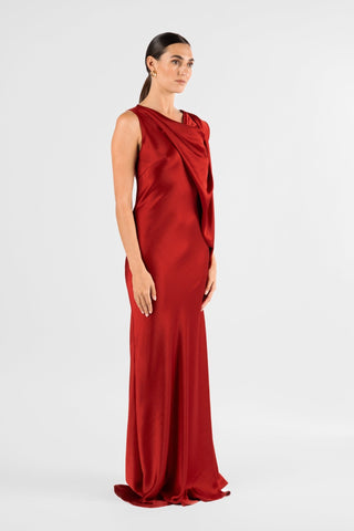 One Fell Swoop Glinch Gown in Carmine, side three-quarter view of the deep red satin gown, showing the cowl neckline, the sleeveless armhole, the draped panel crossing the bodice and the skirt skimming the hip before flaring to a floor-length hem with a slight sweep.