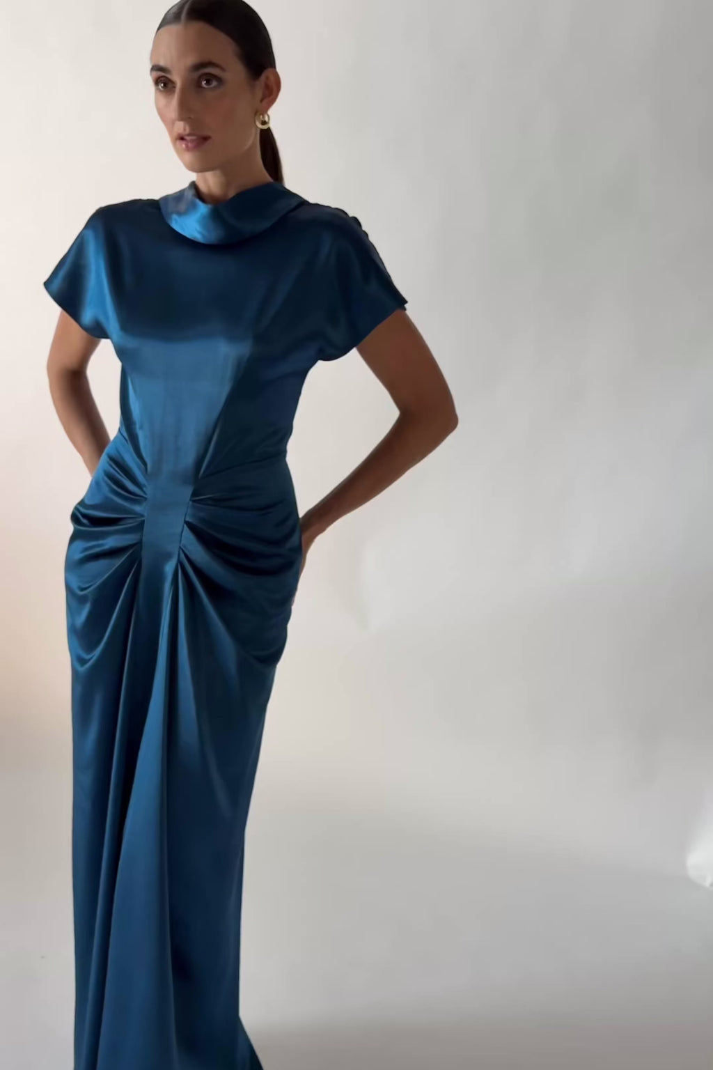 One Fell Swoop Lucent Maxi in Caspian, a video of the model standing front-facing in a lustrous blue satin evening gown with a soft cowl neckline, short sleeves and sculptural ruching through the waist and hips.