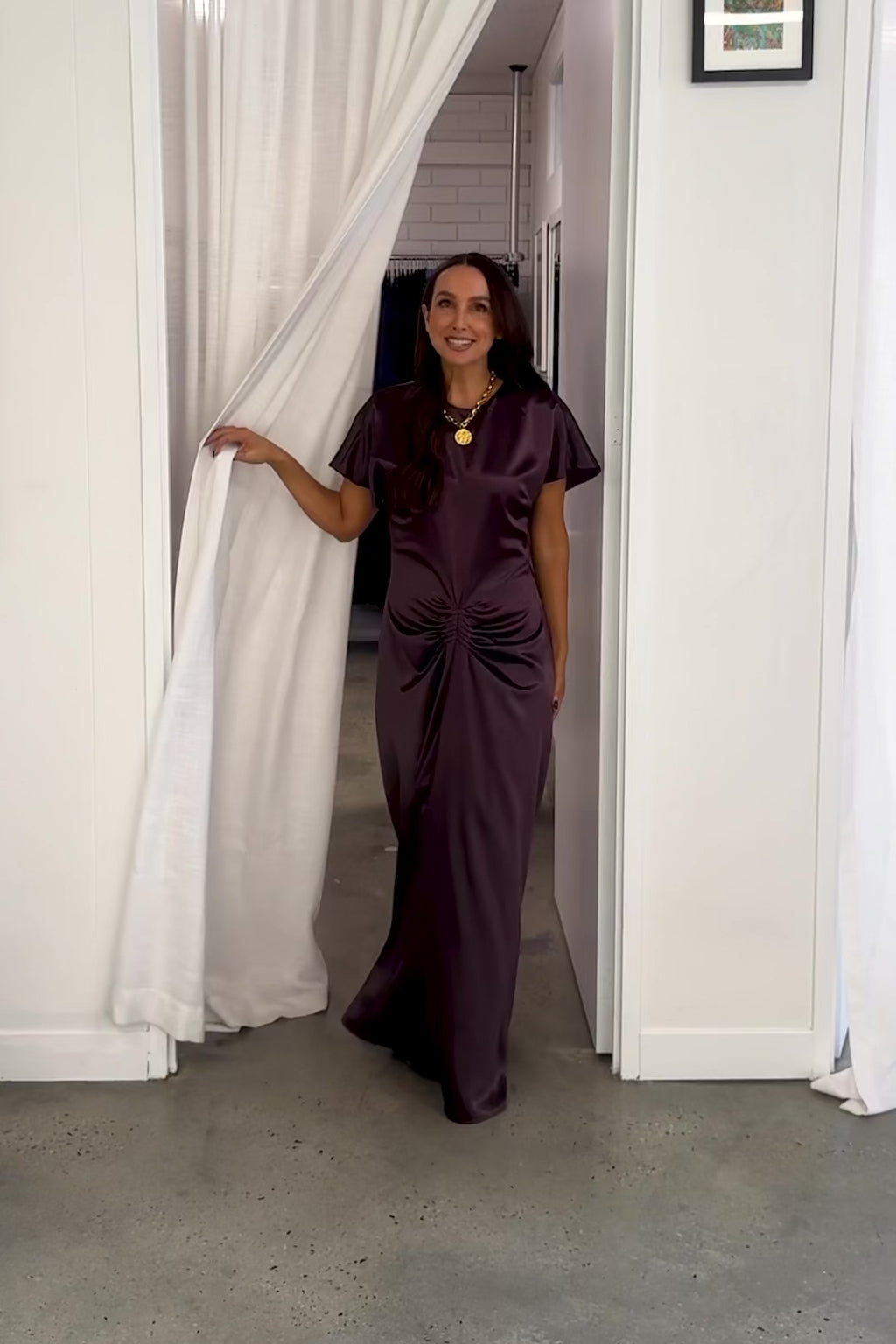 A video showcasing One Fell Swoop's Kiera Dress in Aubergine satin, featuring a bateau neckline, flutter sleeves, central ruched waist and flowing maxi skirt.